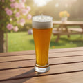 Pepper & White Happy Hour Fressia scented candle in a beer glass shape with white wax foam on a wooden table outdoors.