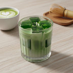 Pepper & White matcha latte candle in a glass with green tea ice cubes, next to a matcha bowl and bamboo whisk.