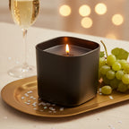 Pepper & White Noir Champagne scented candle in a matte black square vessel on a gold tray with grapes and champagne